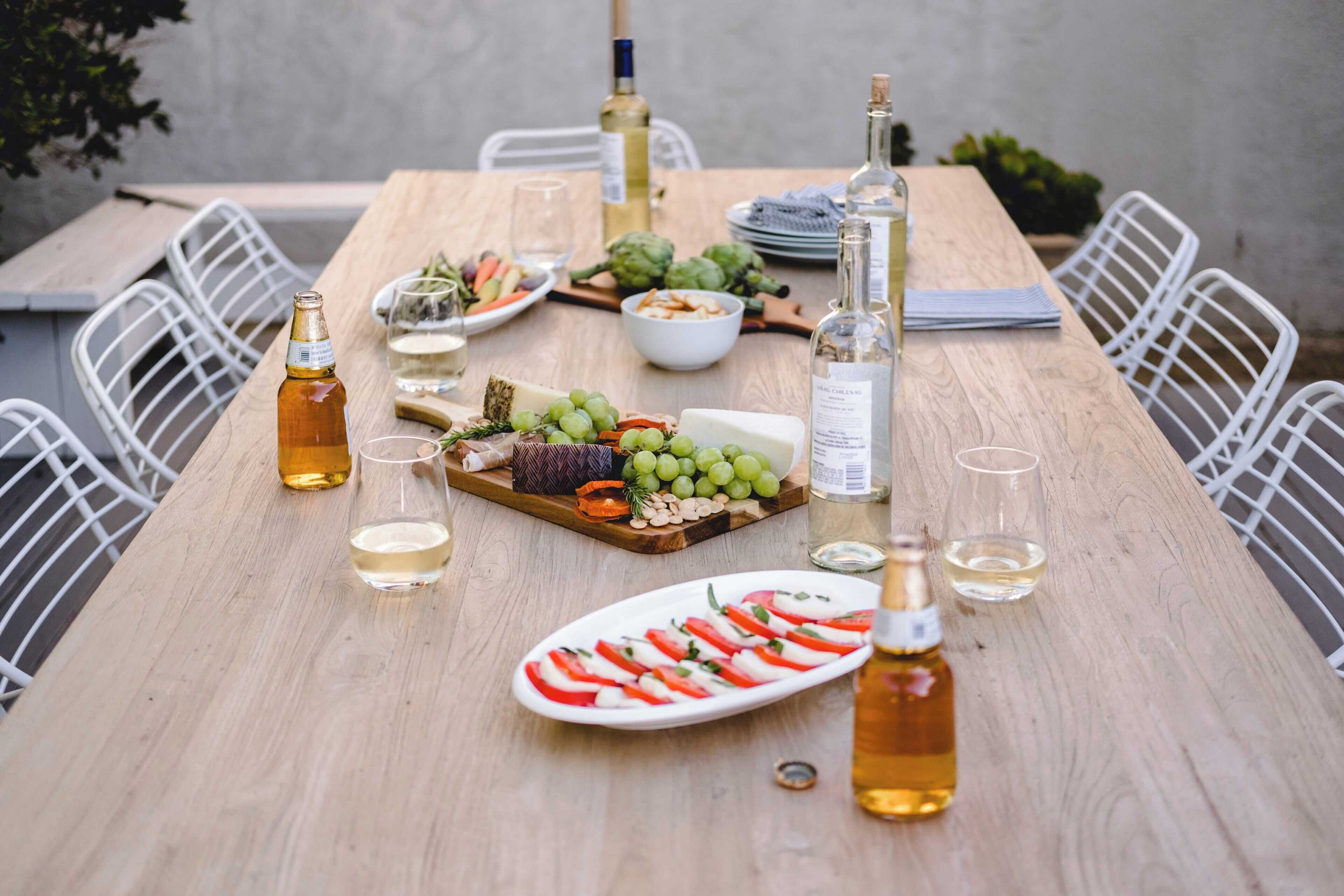 A wooden table set with various food items, bottles, and glasses, surrounded by white chairs.
