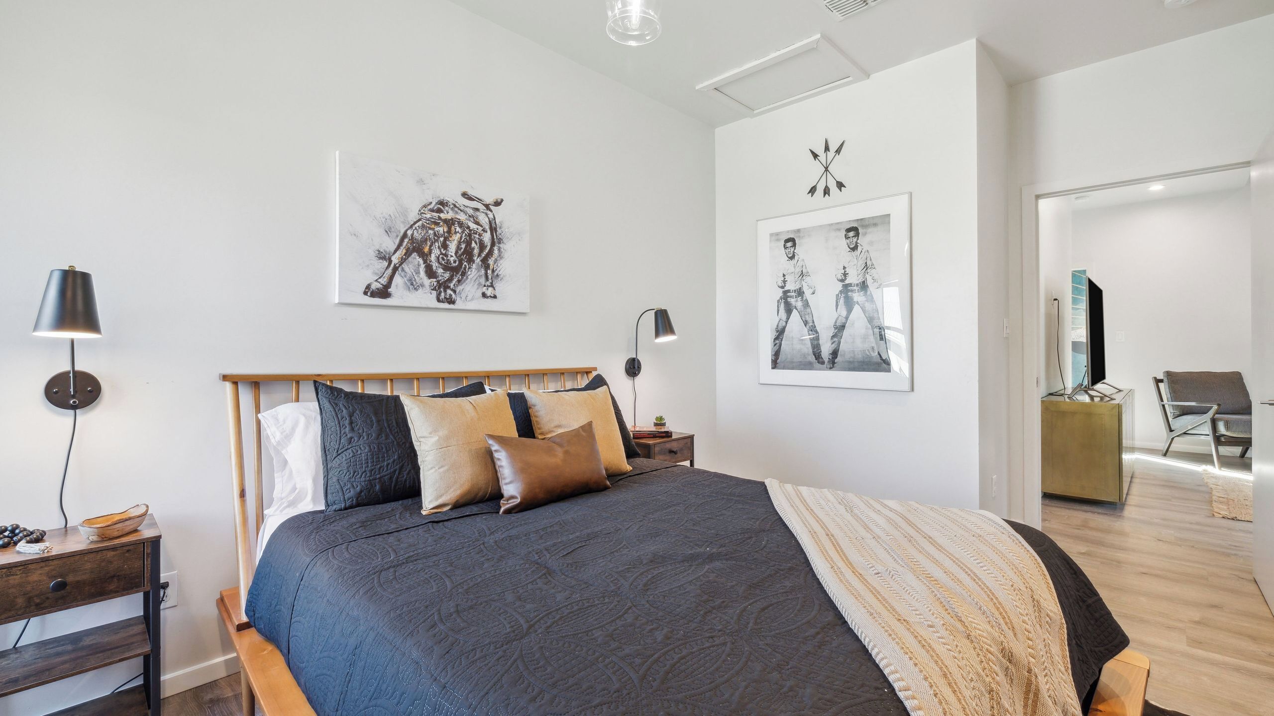 A modern bedroom with a large bed, two bedside tables, a desk, a chair, and artwork on the walls.