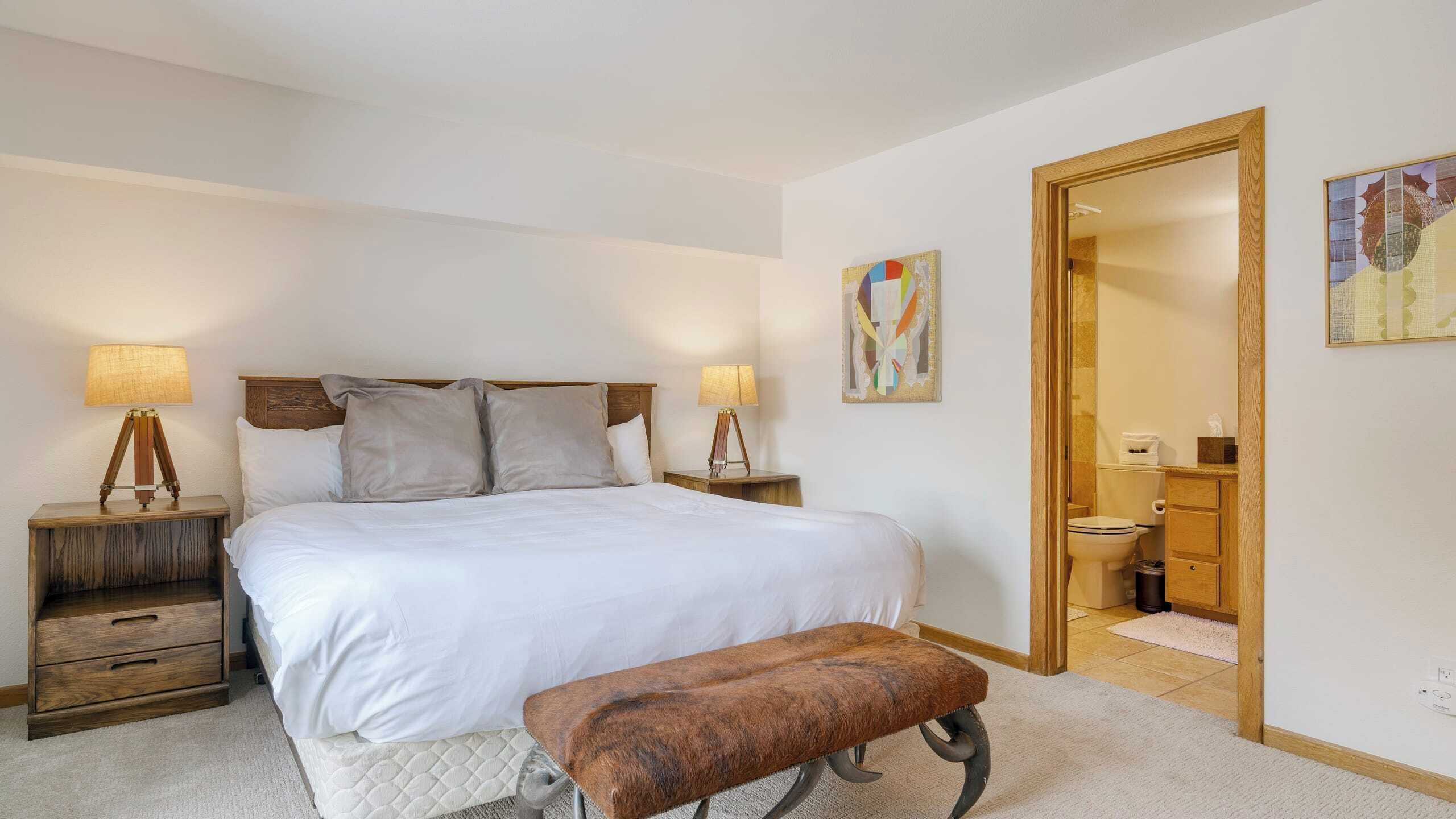 A bedroom with a large bed, two bedside tables with lamps, a bench, and a door leading to a bathroom.