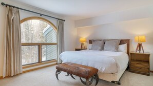 A bedroom with a large window, a bed with white linens, two bedside lamps, a wooden bench, and a small wooden nightstand. - Riverside Condos C102 by Avantstay Condo Close To Downtown, Town Park & Ski Lift #8 (Telluride)