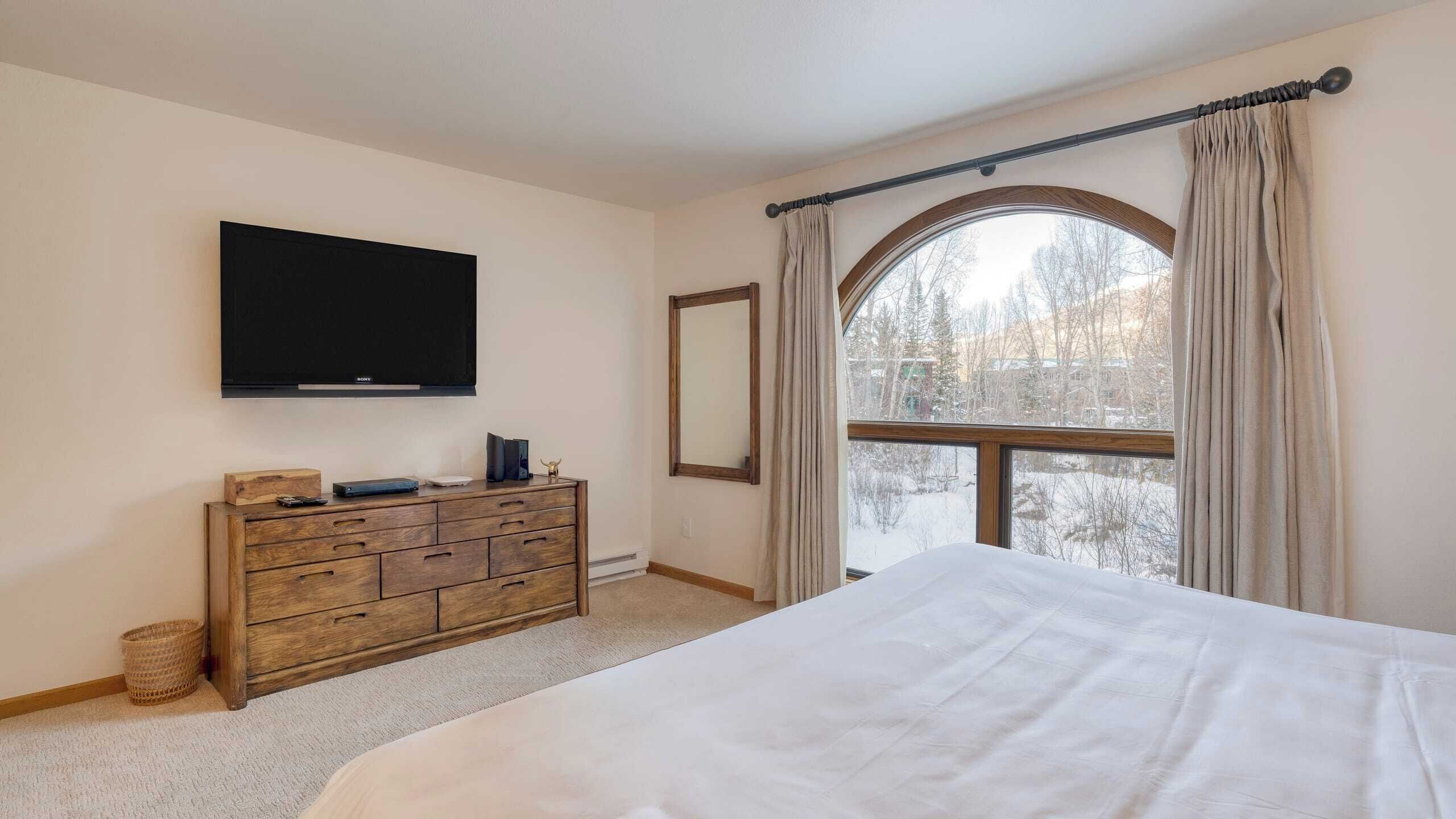 A bedroom with a large bed, a dresser, a flat-screen TV, a window with curtains, and a view of trees outside.