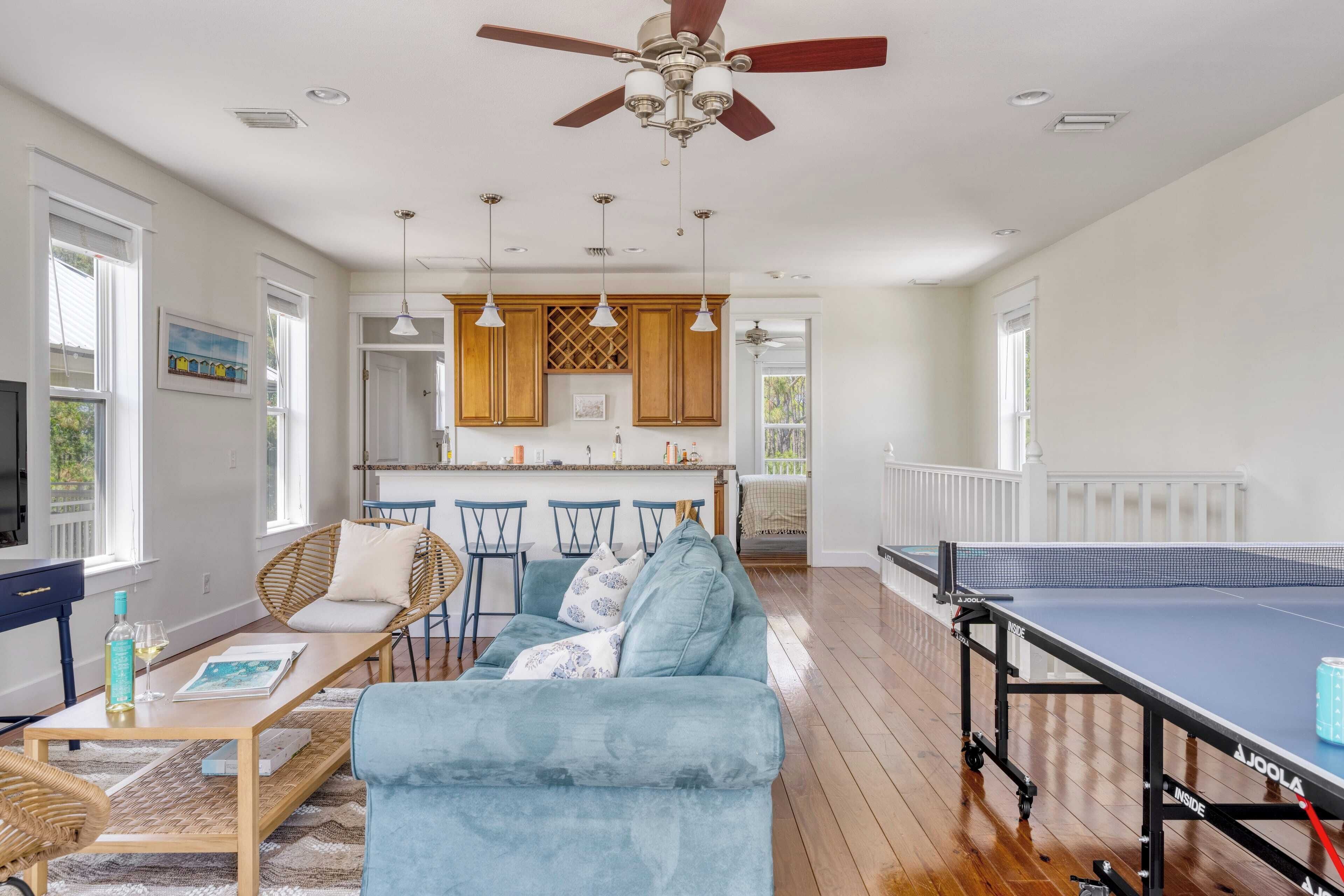 A spacious living room with a ping pong table, a sofa, and a dining area with a kitchen in the background.