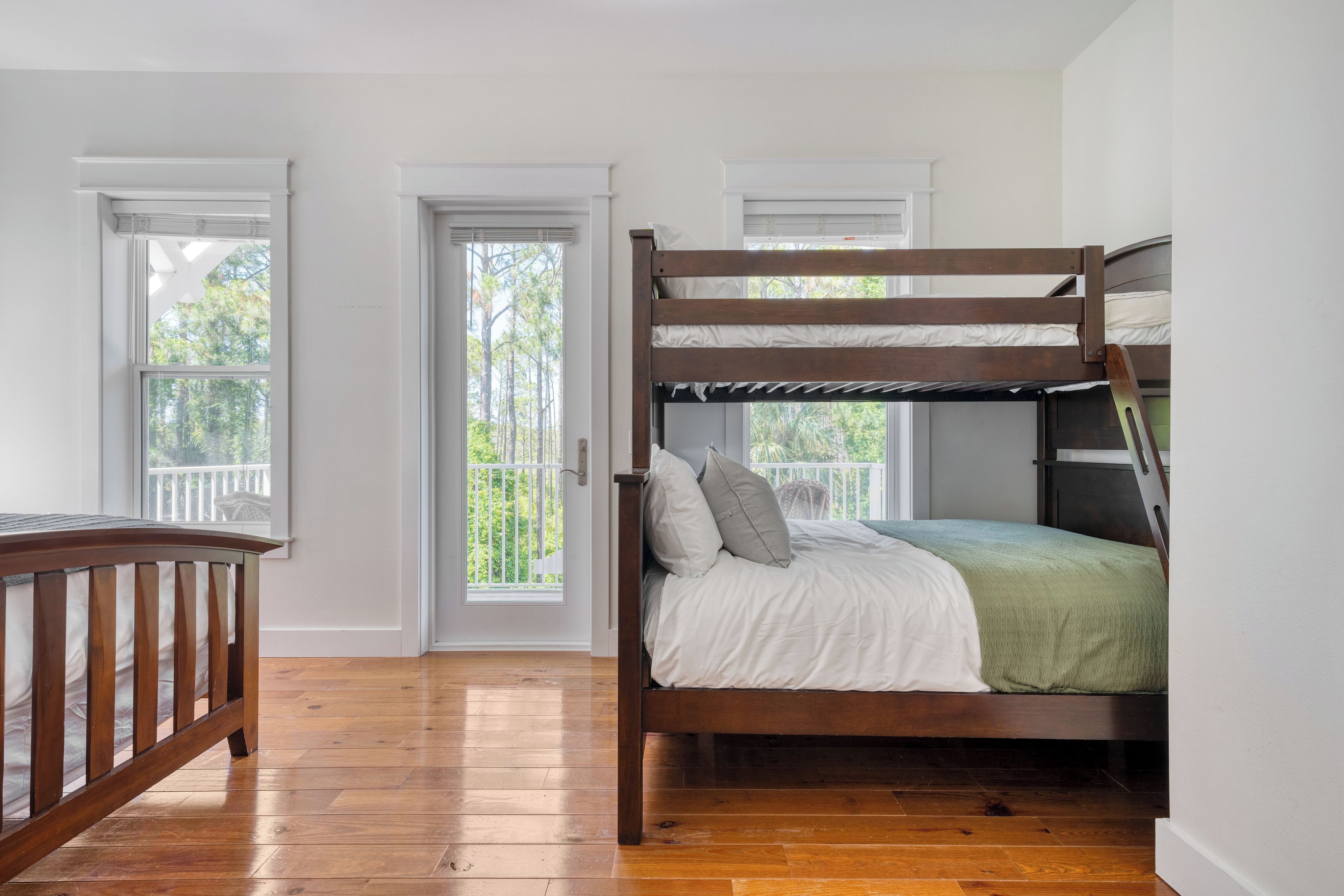 A bunk bed with a wooden frame, a white mattress, and a green blanket. There are two windows with white frames and a door with a white door frame.