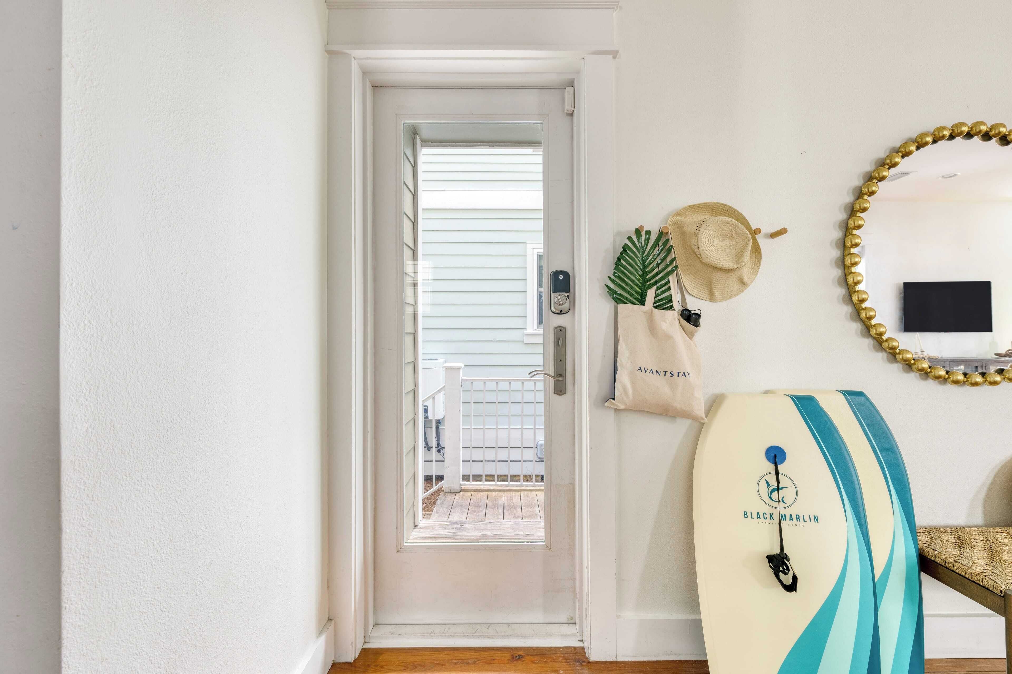 Un couloir avec une porte blanche donnant sur un balcon, une planche de surf, un chapeau et un miroir avec un cadre doré.