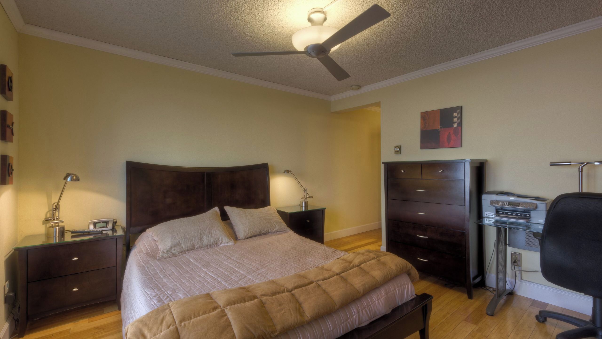 A bedroom with a bed, bedside tables, a desk with a chair, a ceiling fan, and a dresser.