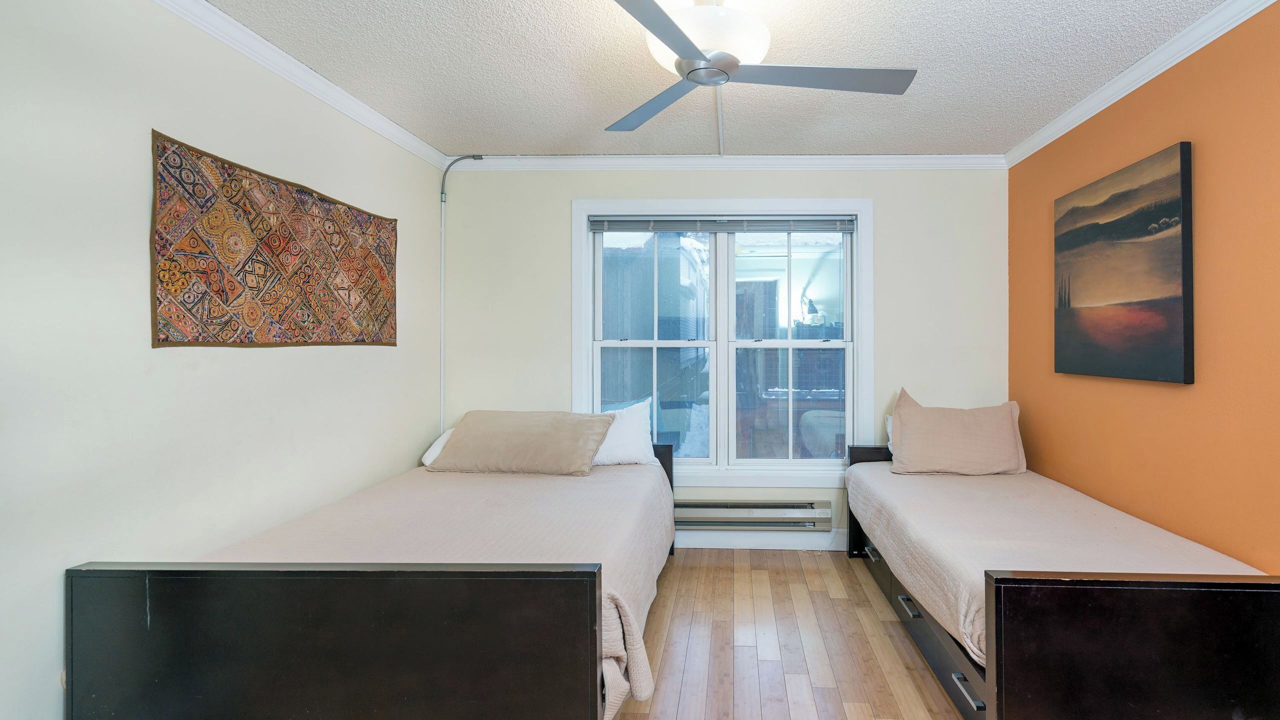 A small room with two beds, a ceiling fan, a window with a view, and a painting on the wall.