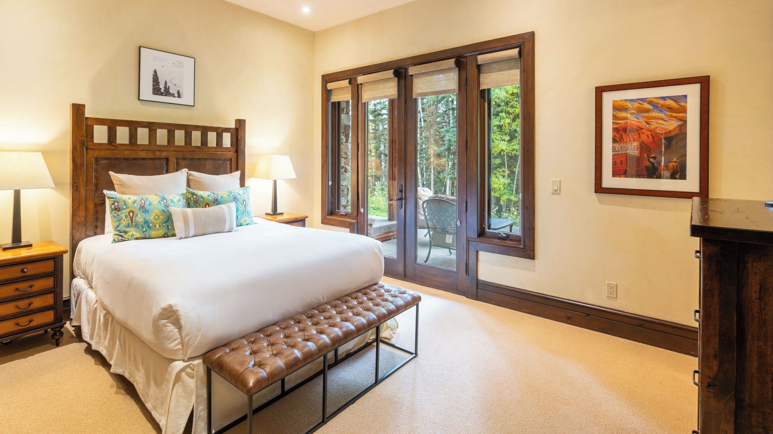 A bedroom with a large bed, a bench, a wooden headboard, two bedside tables with lamps, a framed picture on the wall, and a sliding glass door leading to an outdoor area.
