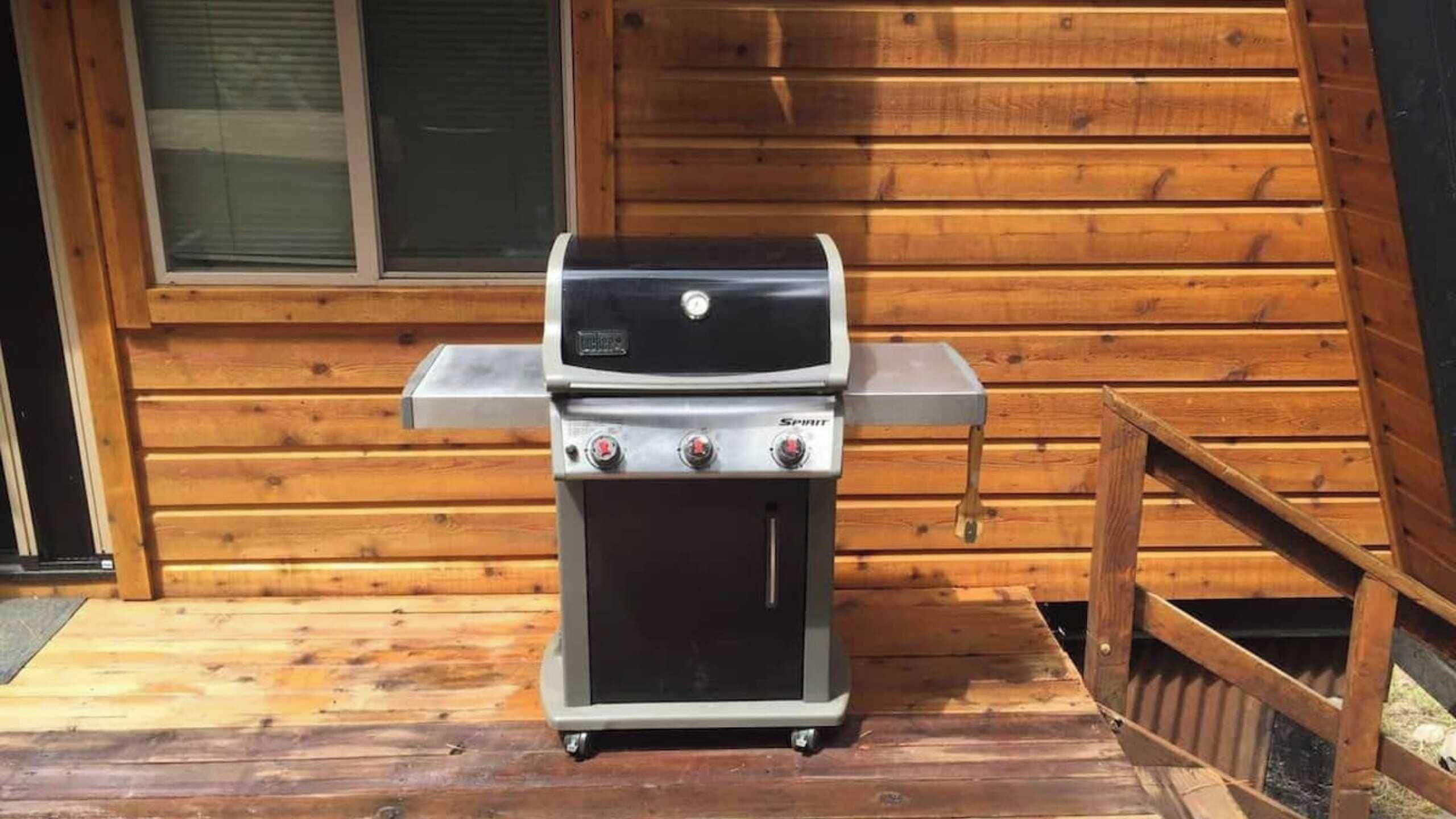 En transportabel Spirit-grill med tre brændere og et digitalt display, placeret på en træterrasse foran en hytte med et vindue og en dør.