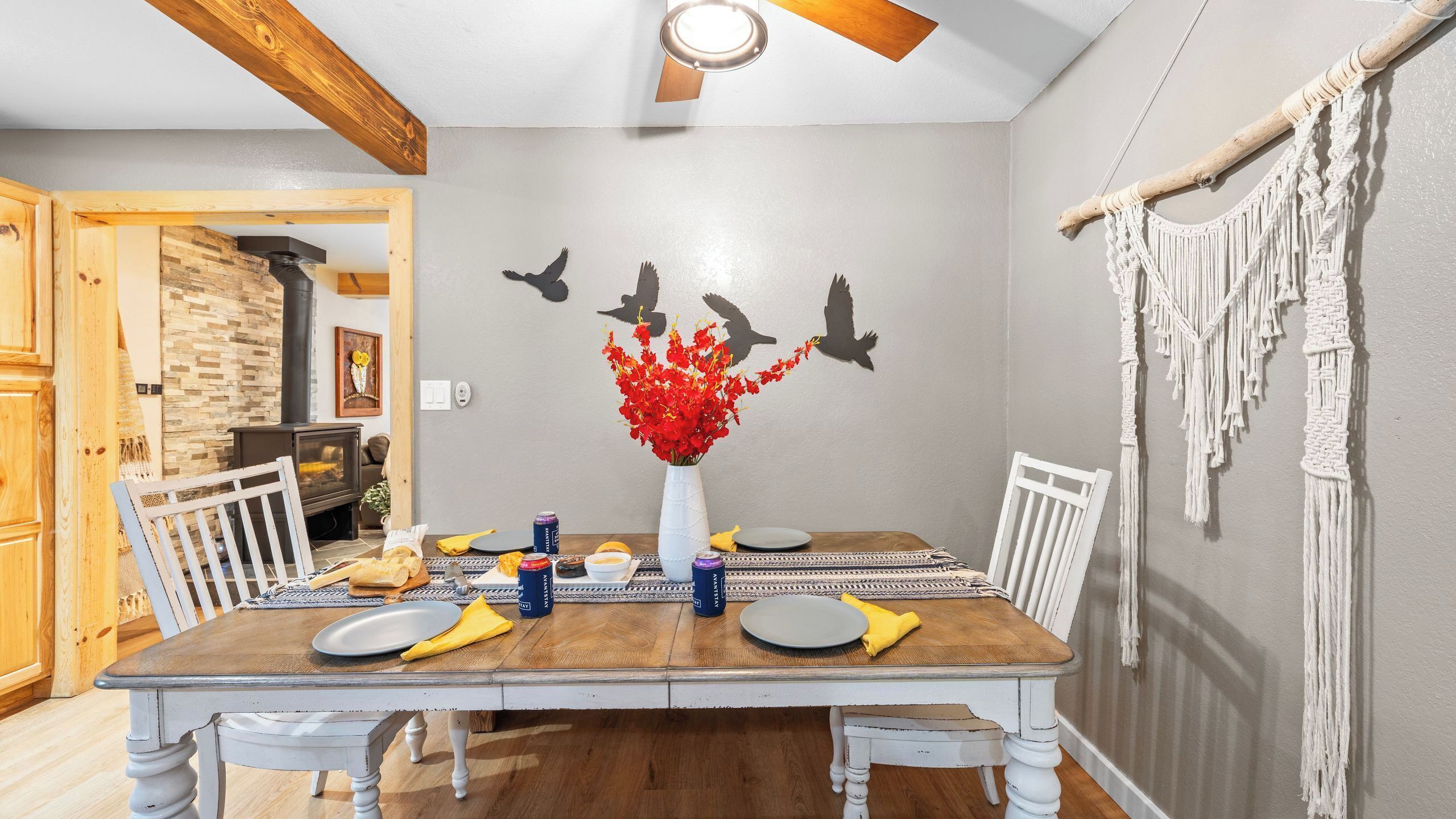 A dining area with a wooden table, white chairs, a vase of red flowers, and wall art of birds.