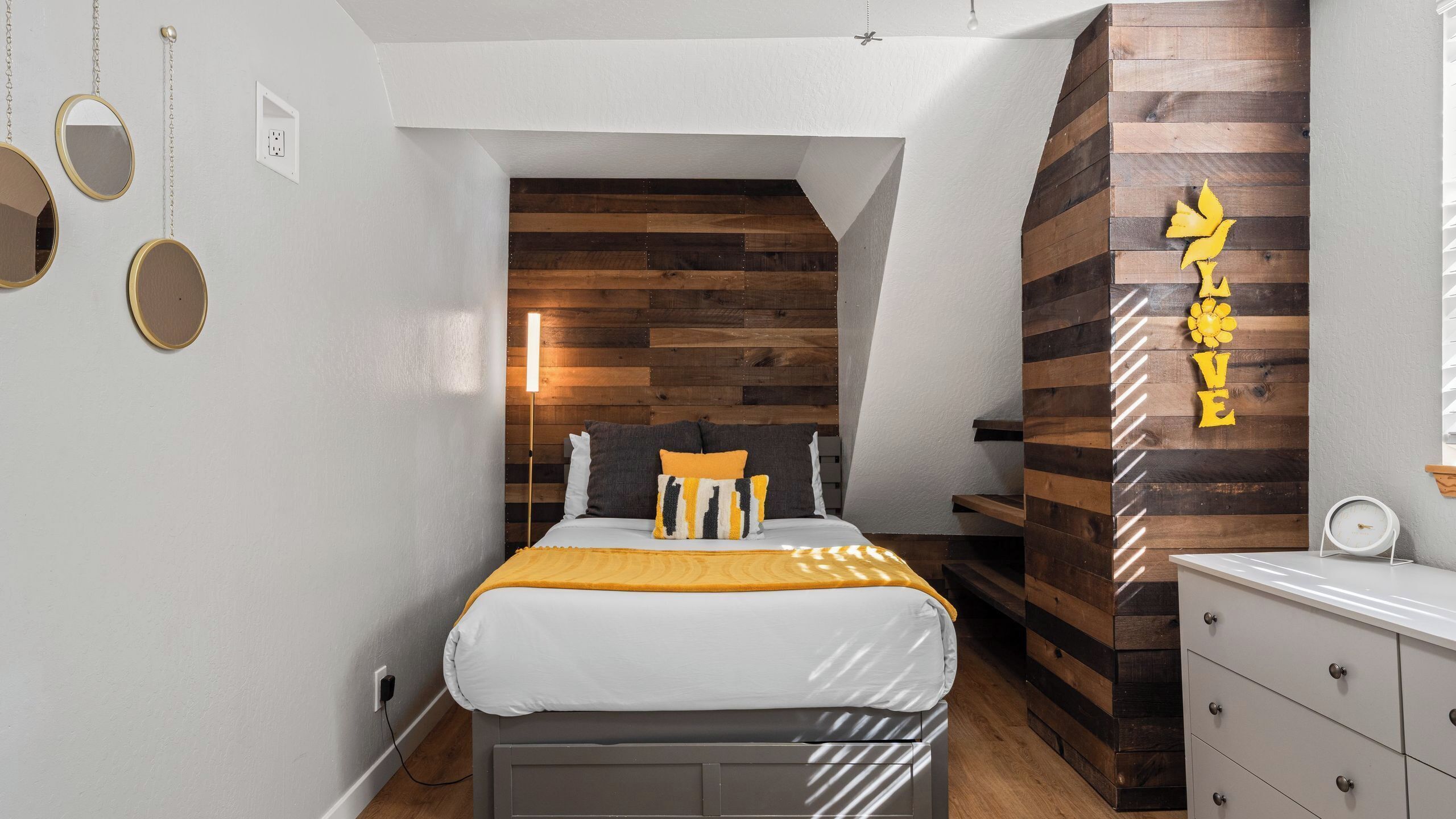 Une chambre à coucher comprenant un lit, une table de chevet, une commode et un mur recouvert de panneaux en bois, sur lequel est accroché un panneau jaune.