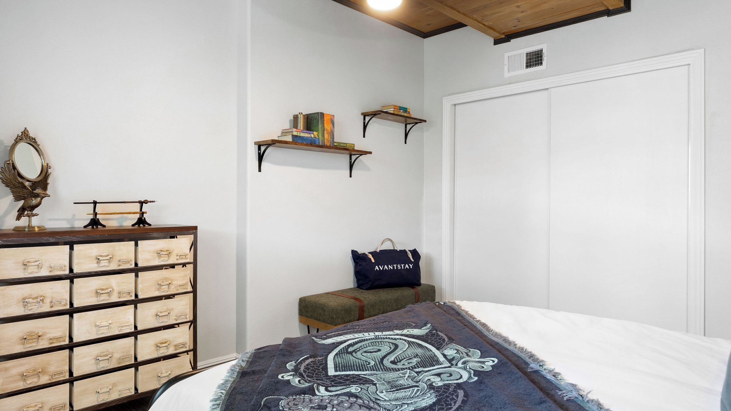 A bedroom with a bed, a dresser with drawers, a mirror, a shelf with books, a bag, and a door.