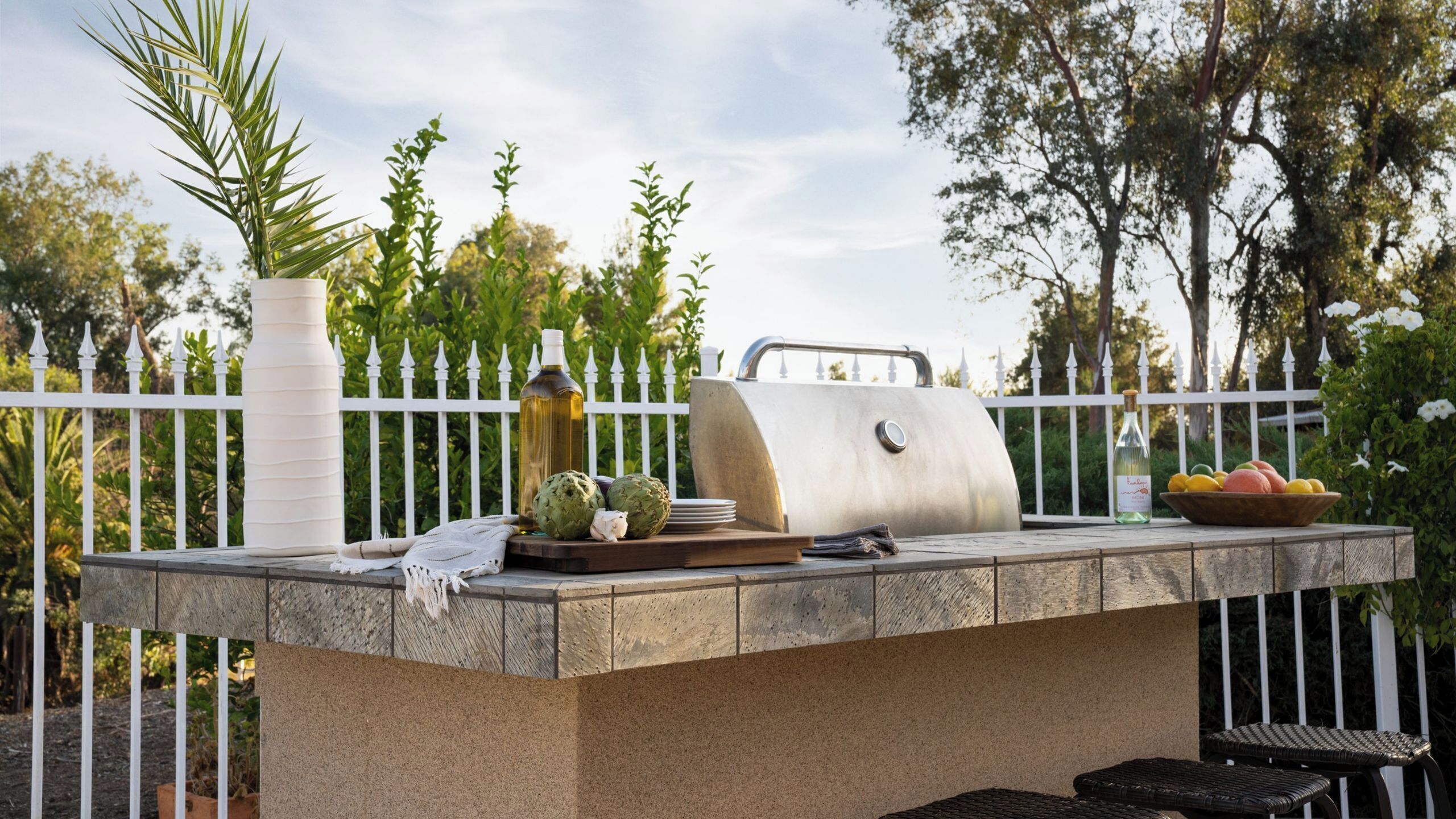 An outdoor grill with a white fence, artichokes, a bottle, and a bowl of fruit.