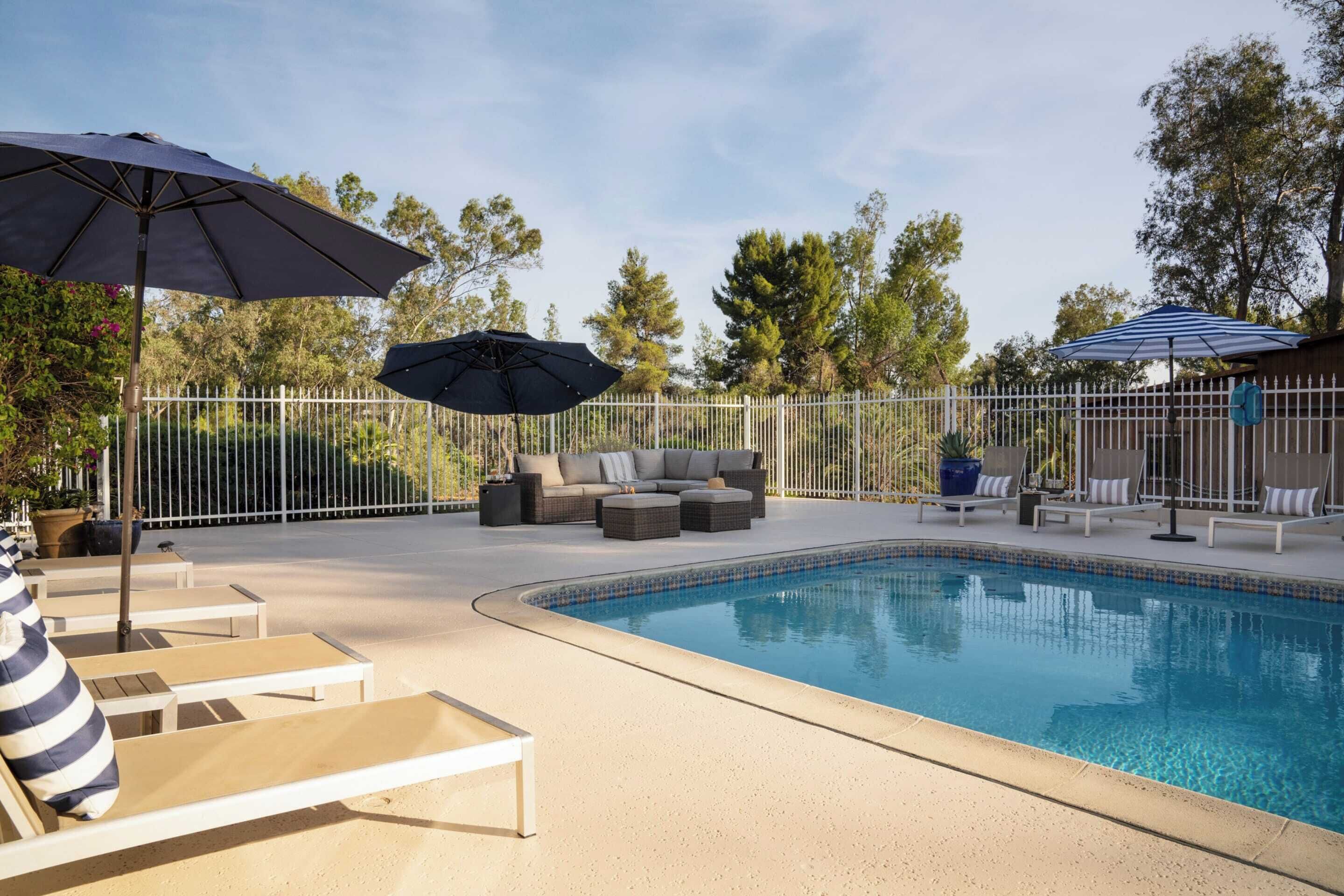 A swimming pool with lounge chairs and umbrellas, surrounded by a fence and trees.