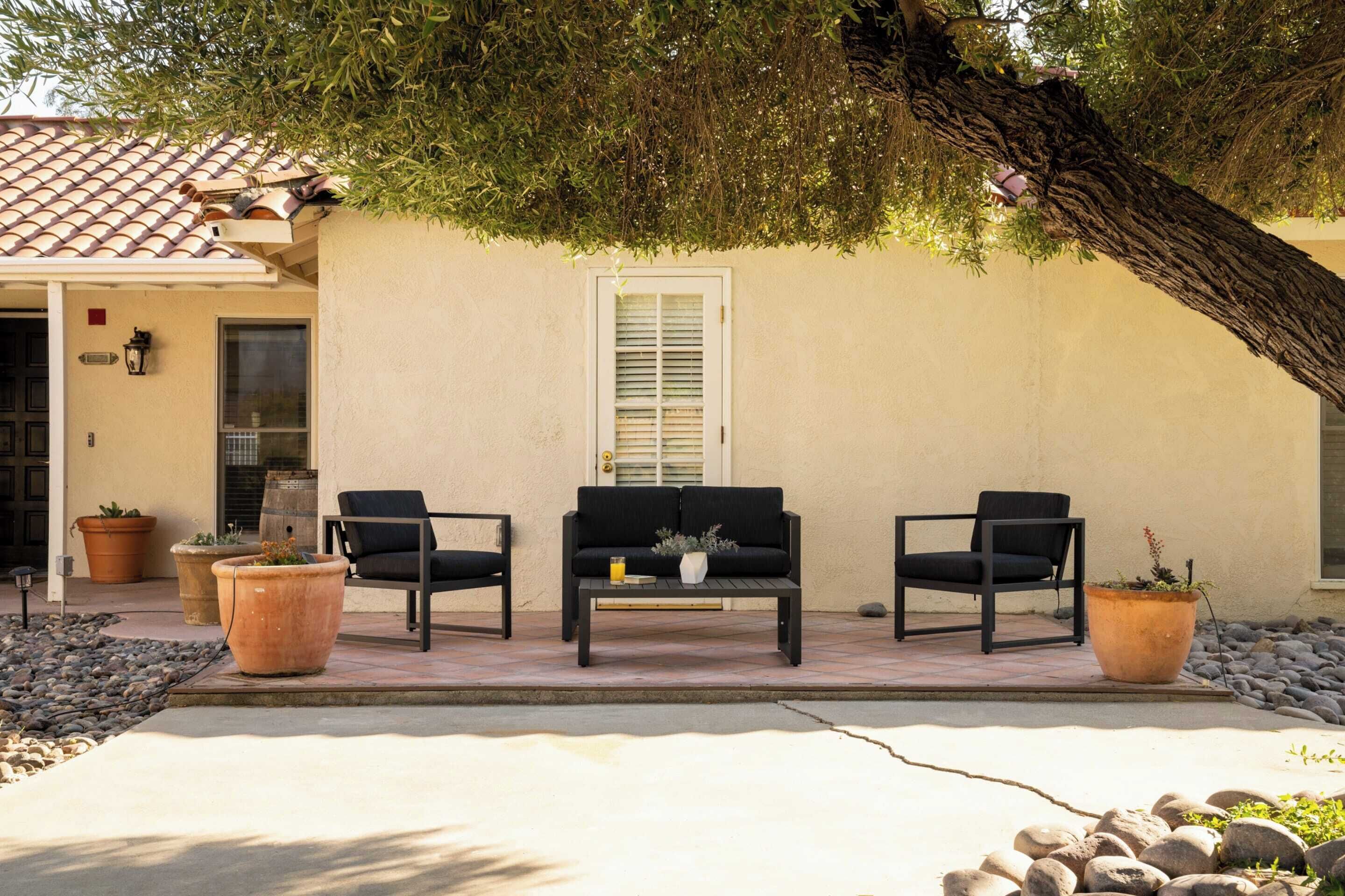 An outdoor seating area with a sofa and two chairs, surrounded by potted plants and a tree.