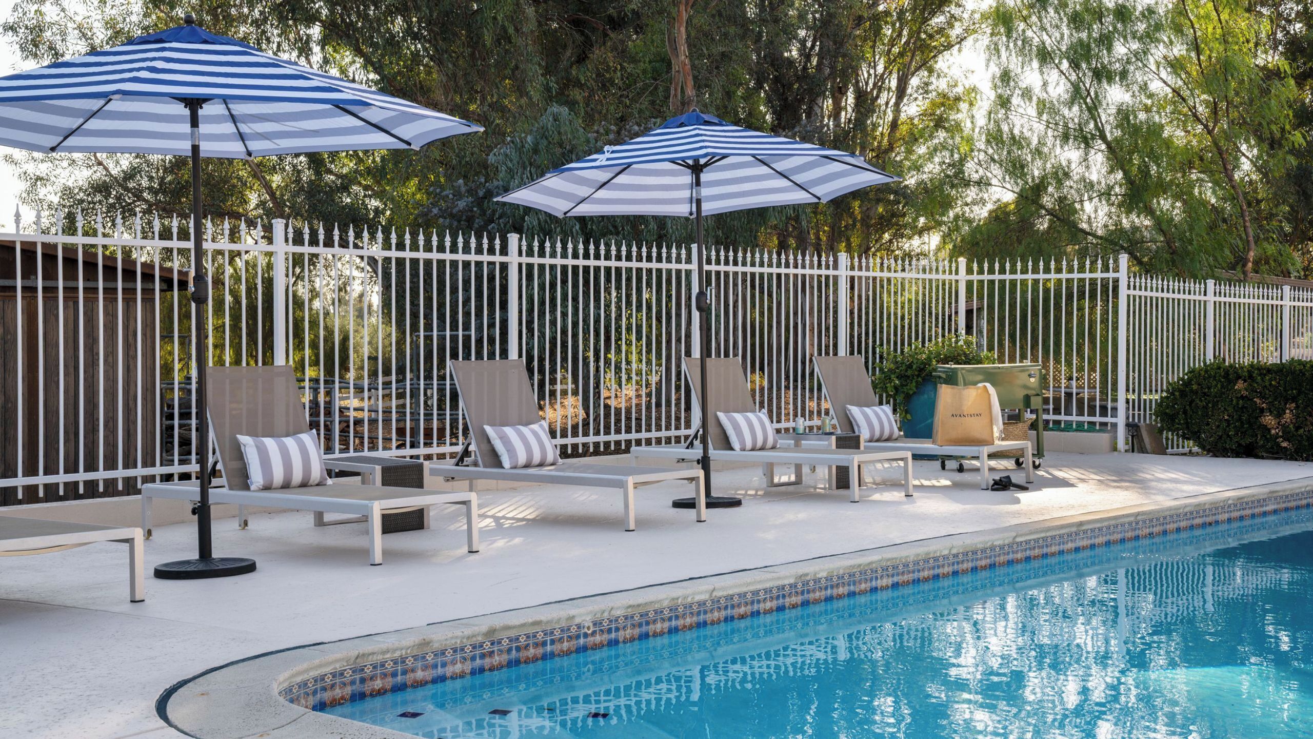 Un espace aménagé au bord de la piscine, avec des chaises longues et des parasols à rayures, entouré d’une clôture blanche et d’arbres.