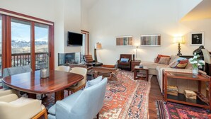 A spacious living room with a large wooden dining table, comfortable seating, a sofa, and a view of mountains through large windows.