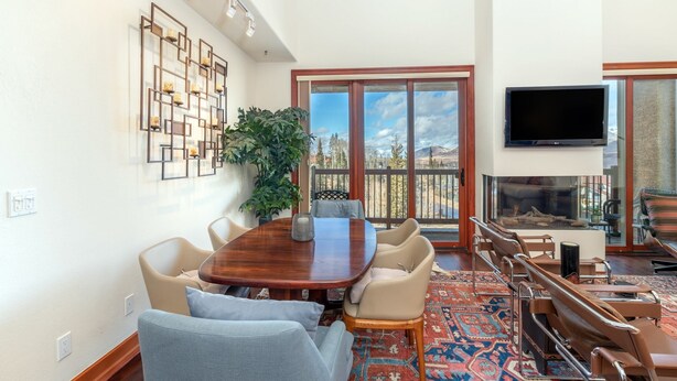 A spacious living room with a large wooden dining table, comfortable seating, a large window with a view, and a flat-screen TV mounted on the wall.