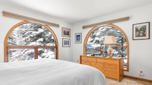 A bedroom with a large bed, wooden dresser, and arched windows overlooking a snowy landscape. - Riverside Condos A103 by Avantstay Condo Close To Downtown, Town Park & Ski Lift #8 (Telluride)
