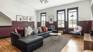 Un salon moderne avec un canapé d’angle, une table basse ronde et un tapis à motifs. Il y a deux fenêtres équipées de stores, un luminaire au plafond et des œuvres d’art encadrées accrochées aux murs.