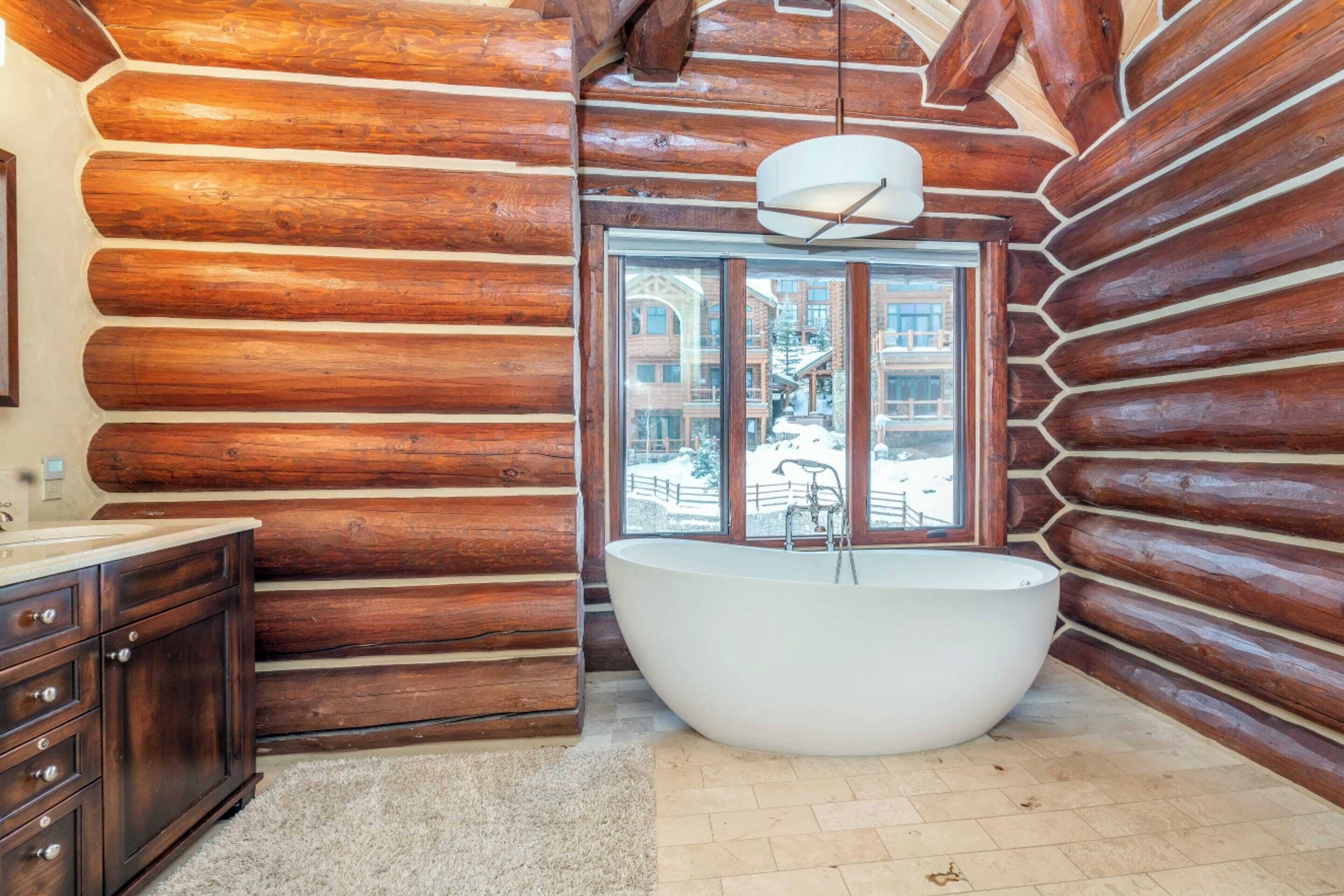 A log cabin bathroom with a large bathtub, wooden walls, and a window offering a view of a snowy landscape.