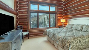 A log cabin bedroom with a large window, a bed, a TV, and a dresser.