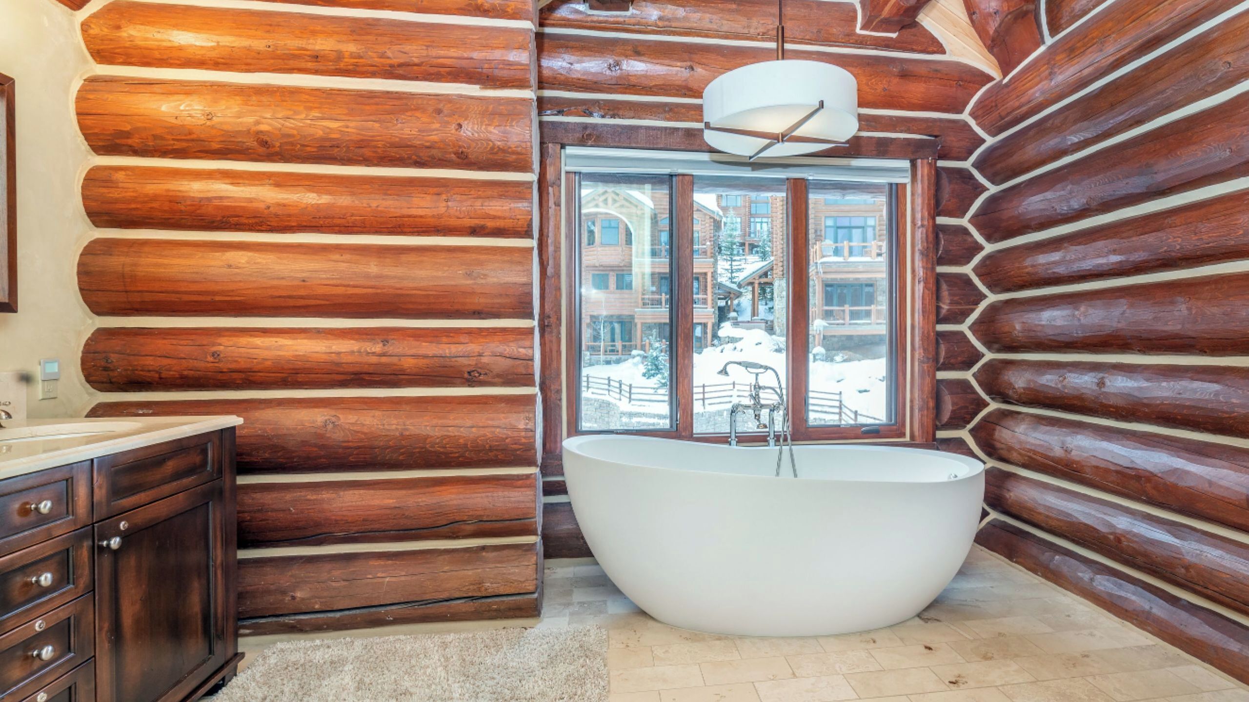 A log cabin bathroom with a large bathtub, wooden walls, and a window with a view of a snowy landscape.