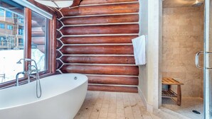 A log cabin bathroom with a freestanding bathtub, a shower, and a window overlooking a snowy landscape.