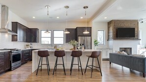 A modern kitchen with dark cabinets, a stainless steel refrigerator, a large island with stools, and a flat-screen TV mounted above the fireplace.