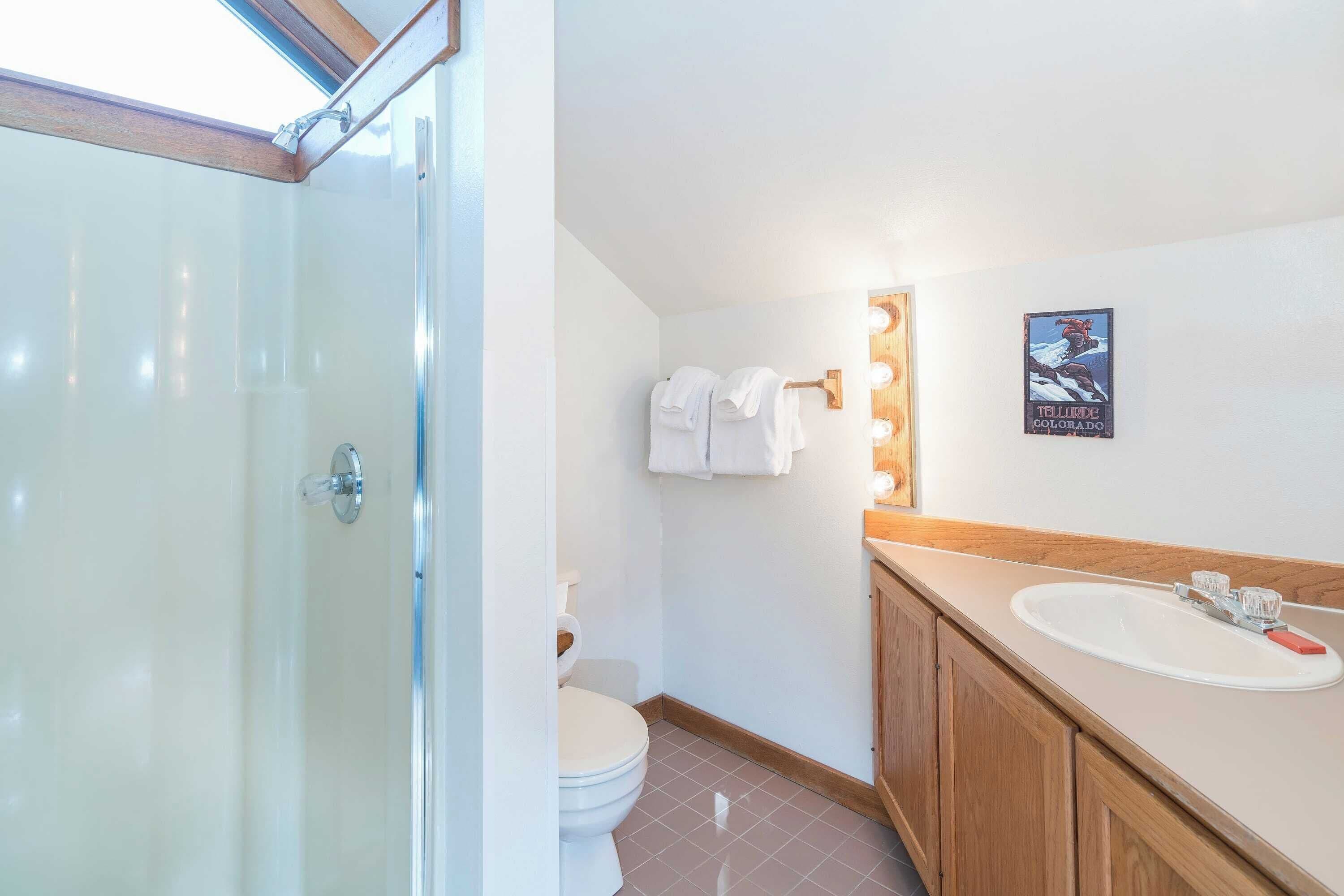 Un baño con mampara de vidrio, inodoro, lavabo con espejo y un cartel en la pared.