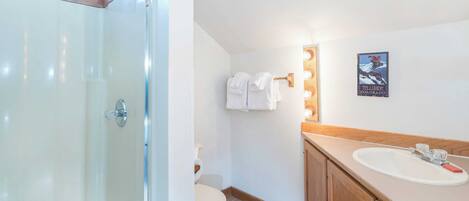 Un baño con mampara de vidrio, inodoro, lavabo con espejo y un cartel en la pared.