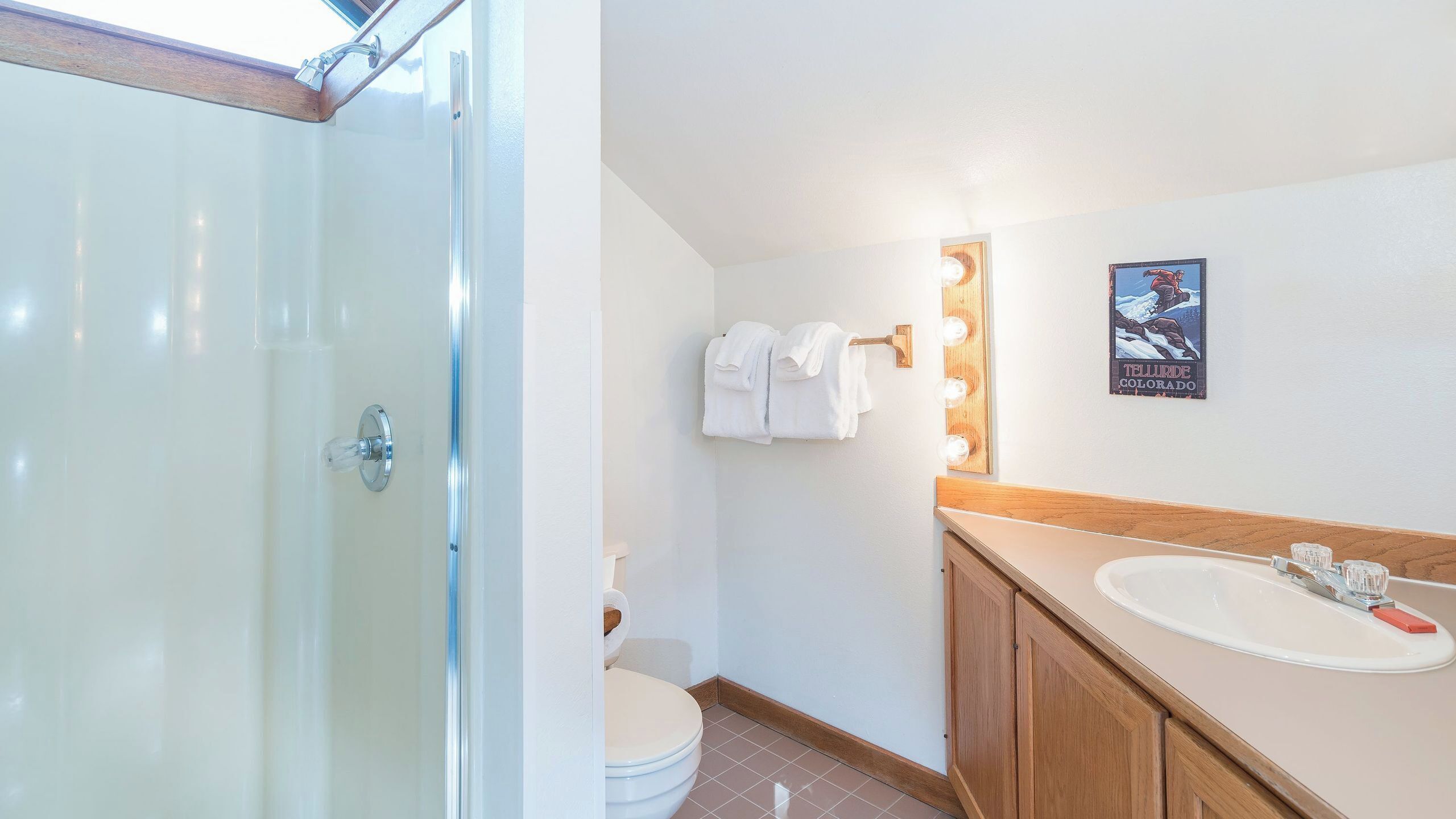 A bathroom with a glass shower door, a toilet, a sink with a wooden vanity, and a poster on the wall.
