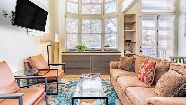 A living room with a sofa, armchairs, a coffee table, and a TV mounted on the wall. Large windows provide natural light and a view of the outdoors. - Columbia Place 5 by Avantstay Ski-in/ski-out Unit in Great Location! (Telluride)