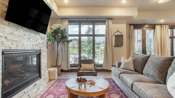 A living room with a fireplace, a sofa, a coffee table with pinecones, and large windows with curtains.