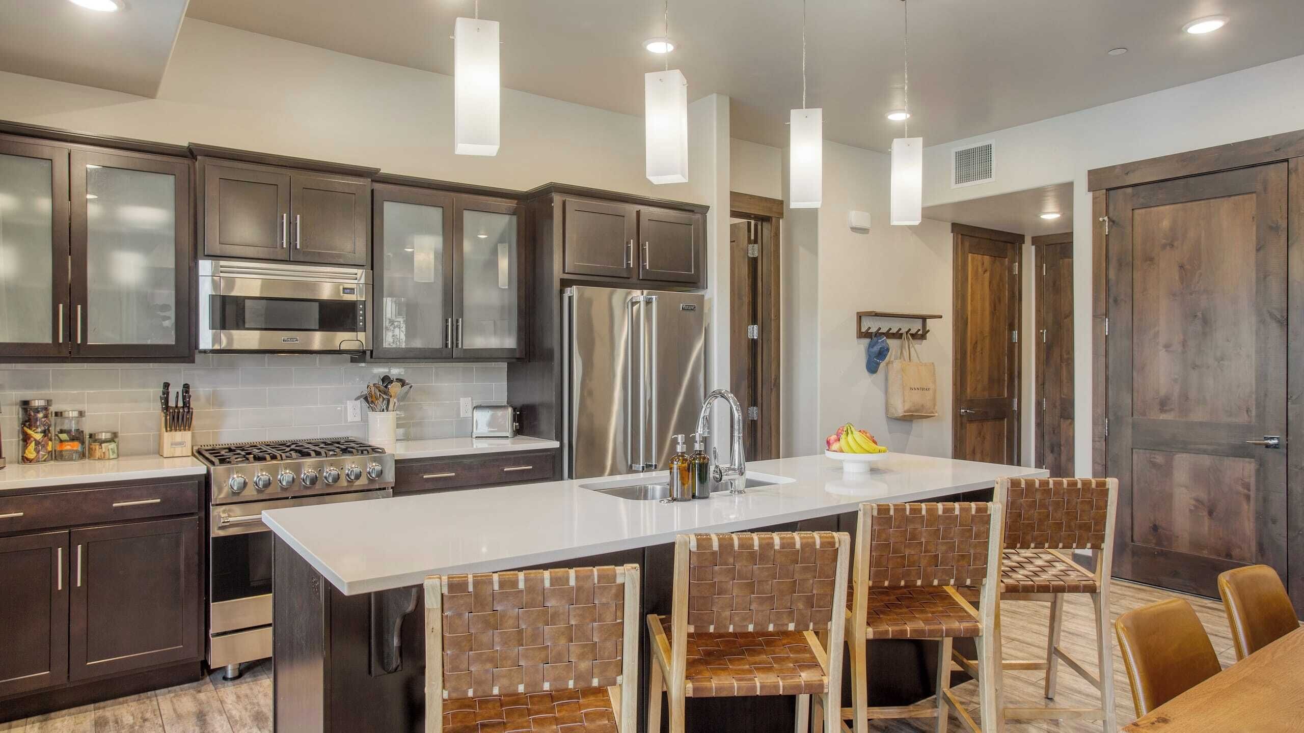 Una cocina moderna con armarios de madera oscura, electrodomésticos de acero inoxidable, una isla central con taburetes altos y luminarias colgantes.