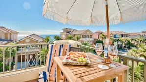 Une table en bois sur laquelle sont disposés un plateau de fruits et des verres à vin, un parasol, et une vue sur un quartier situé sur la côte.