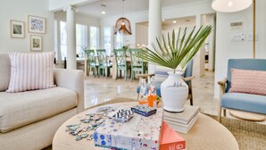 Un salon moderne avec une table basse ronde, un canapé et un fauteuil bleu. Des tableaux encadrés ornent le mur, ainsi qu’un grand vase blanc contenant une plante. On aperçoit également un coin repas au fond de la pièce.
