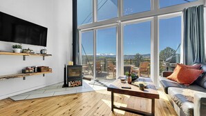 A modern living room with a large window, wooden flooring, a flat-screen TV, a fireplace, and a cozy seating area. - Off-grid Masterpiece Cabin W/ Breathtaking View (Central City)