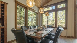 A dining area with a wooden table, leather chairs, and pendant lights. Large windows offer a view of trees. - Sawtooth by Avantstay Spacious Pine Tree Cabin w/ Hot Tub (Truckee)