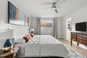 Relax in this serene bedroom with a large bed, nightstand, and dresser, featuring a ceiling fan and mounted TV.