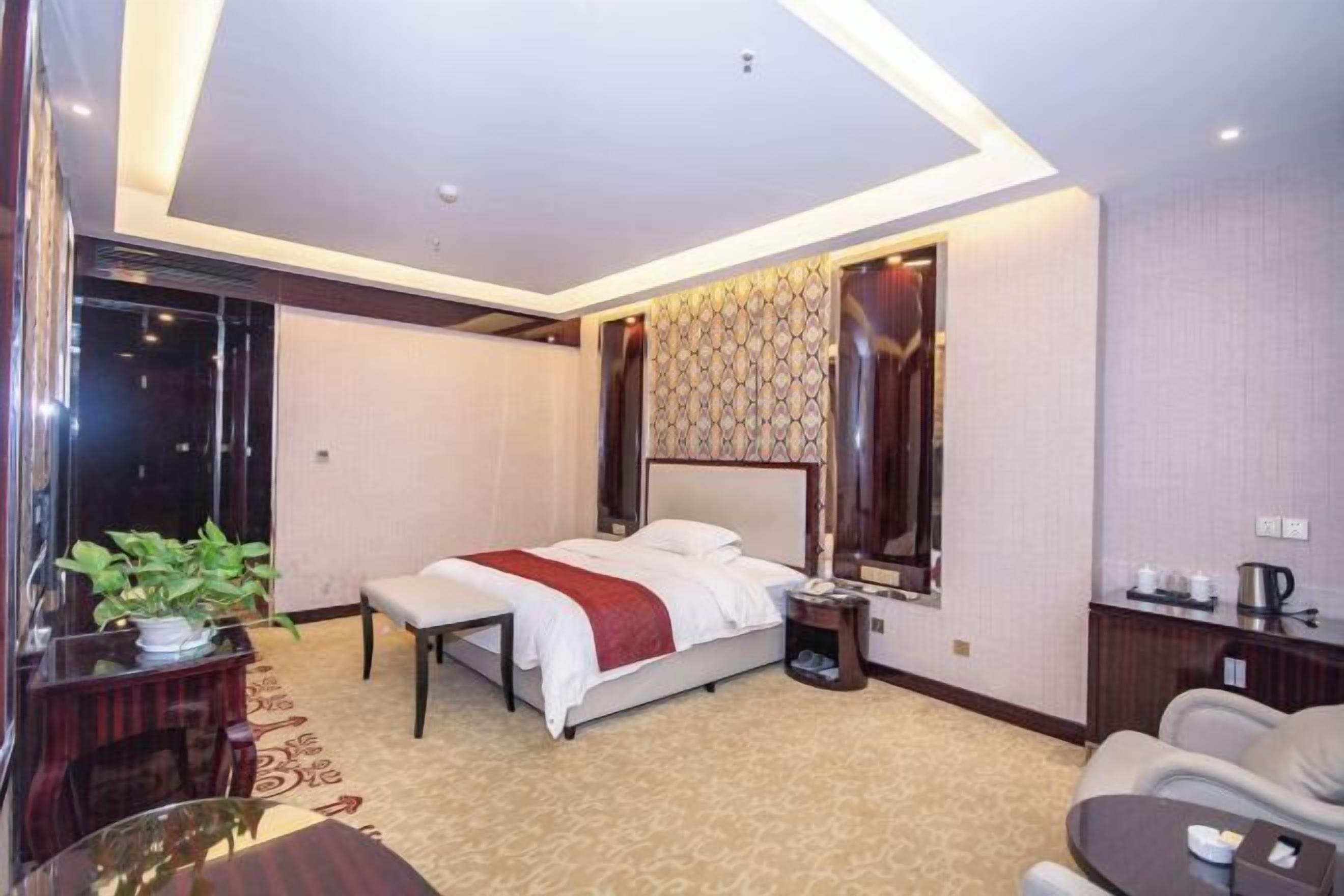 A hotel room with a bed, bedside table, lamp, chair, and a small table with a plant.