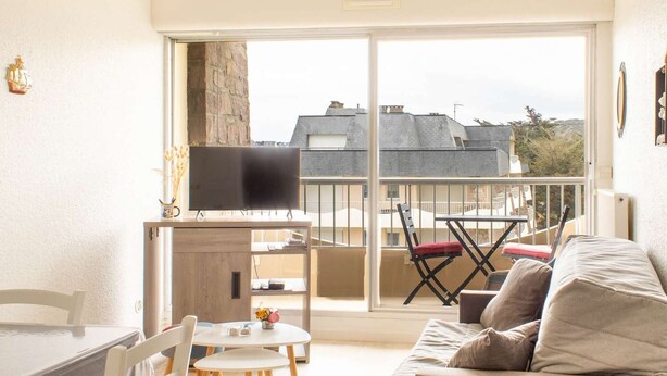 A living room with a sofa, a dining table, and chairs. There is a TV on a stand, and a balcony with a view of buildings and trees. - Sea-view apartment with two balconies in Erquy | 4 guests | Ref 442 (ERQUY)