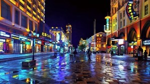 Una calle iluminada de noche, con edificios brillantes y pavimento mojado que refleja las luces.