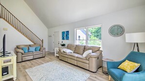 Ein Wohnzimmer mit einem beigefarbenen Sofa, einem blauen Sessel, einer Holztreppe, einer weißen Tür und einem Fenster mit Blick auf Grünflächen.