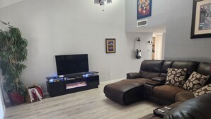 Ein Wohnzimmer mit Ecksofa, einem Flachbildfernseher auf einer schwarzen Unterhaltungsecke, einem Blumentopf und einem Kamin.