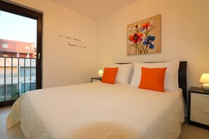A bedroom with a large bed, two nightstands, a window with a view of buildings, and a wall with a floral painting and inspirational quote.