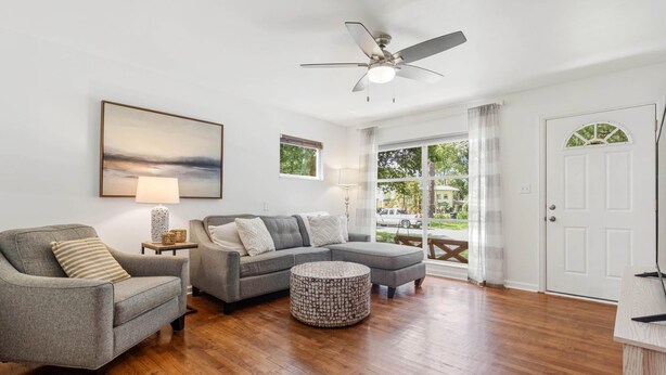 A living room with a grey sofa, a grey armchair, a round ottoman, a ceiling fan, and a large window with a view of greenery.