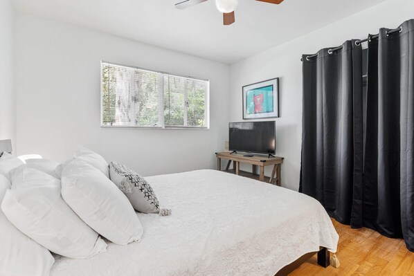 Une chambre à coucher avec un lit, un téléviseur posé sur un meuble en bois, une fenêtre avec des stores et un rideau noir.