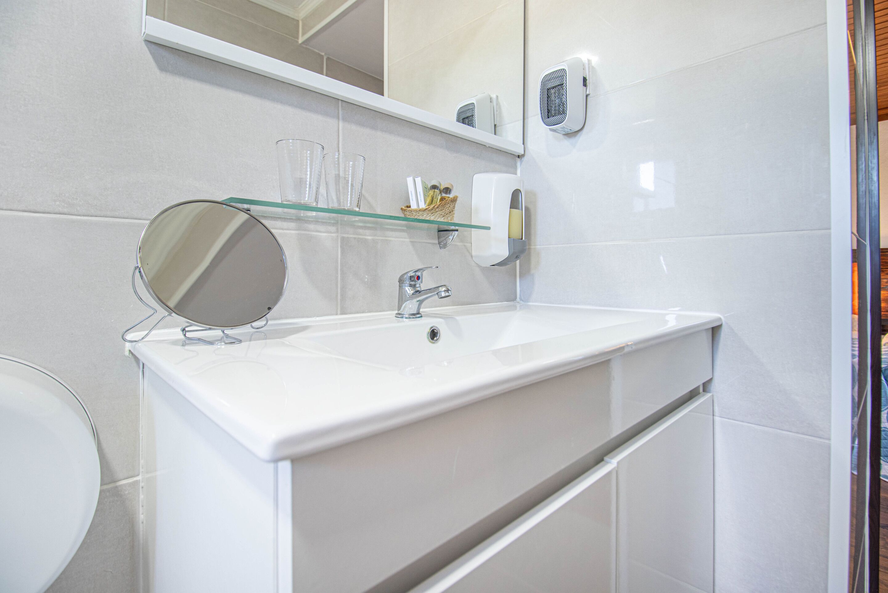 Un baño con un lavabo blanco, un espejo, un estante de vidrio con artículos de tocador y un dispensador de toallas.