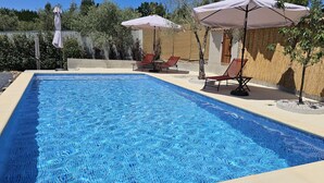 A rectangular swimming pool with clear blue water, surrounded by a tiled deck. There are two red lounge chairs and two umbrellas providing shade. - Quinta Penedo das Antas (Serta)