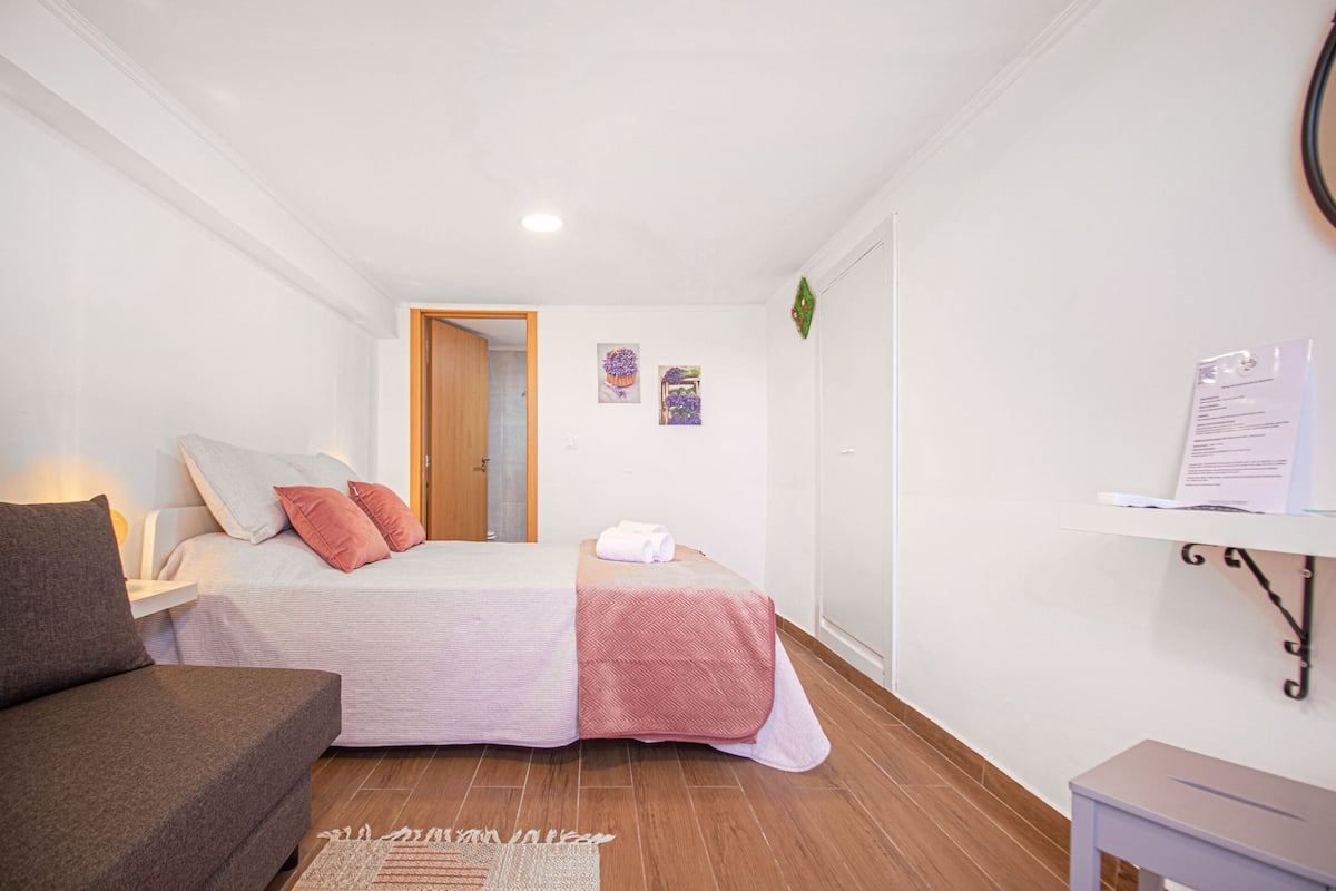 A small, well-lit bedroom with a bed, a sofa, a small table, and a wall-mounted shelf.